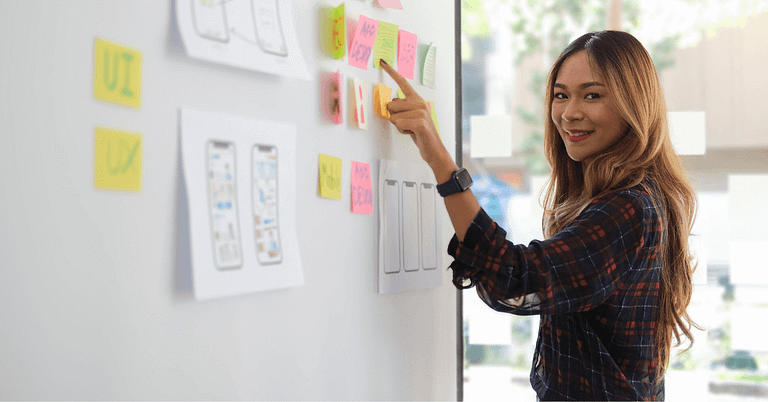 Asian woman pointing at sticky notes related to JavaScript frameworks on a wall.