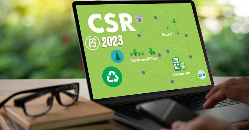 A laptop screen displays a presentation on corporate social responsibility (CSR) for the year 2023, highlighting full-scale outreach programs and corporate responsibility themes, with a pair of glasses and a smartphone positioned on
