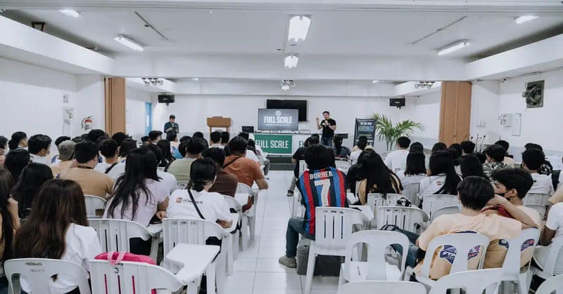 A speaker presents a career talk to an audience in a conference hall or seminar room, with attendees seated facing the stage.