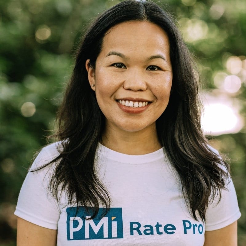 Asian woman smiling at the camera, wearing a white t-shirt with "pmi rate pro" logo, standing against a blurred greenery background.