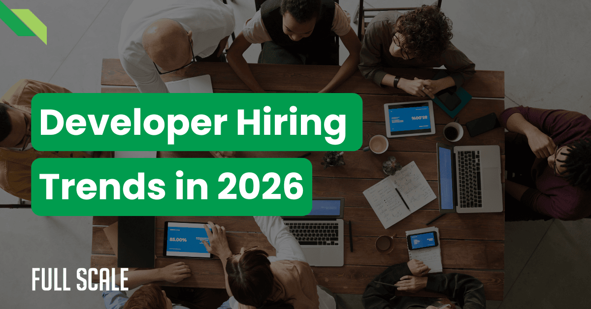A group of people with laptops and tablets sit around a wooden table; text overlay reads “Developer Hiring Trends in 2026”—highlighting the software developer shortage 2026.