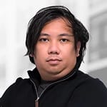 A person with medium-length dark hair, wearing a black jacket, looking at the camera with a neutral expression. The background is blurred.