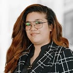 A person with long, wavy hair and glasses is wearing a plaid blazer and black blouse, standing against a blurred background.