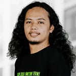 A person with long, curly hair and a beard is standing indoors, wearing a black T-shirt and facing the camera with a neutral expression.