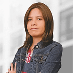 A person with long brown hair wearing a denim jacket and a striped shirt stands with arms crossed in front of a blurred urban background.