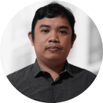 A man with short black hair wearing a dark button-up shirt, facing the camera with a neutral expression against a blurred light background.