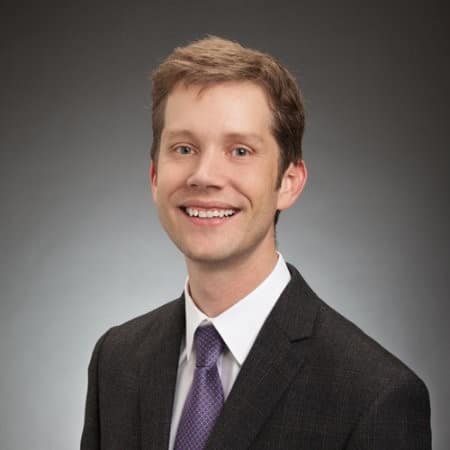 Smiling man with short brown hair wearing a dark suit, white shirt, and lavender tie against a gray background.