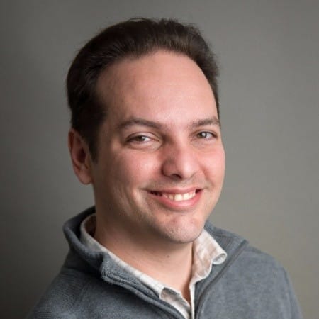 A man with short dark hair smiling, wearing a light gray quarter-zip sweater and a light-colored collared shirt, posing against a plain gray background.