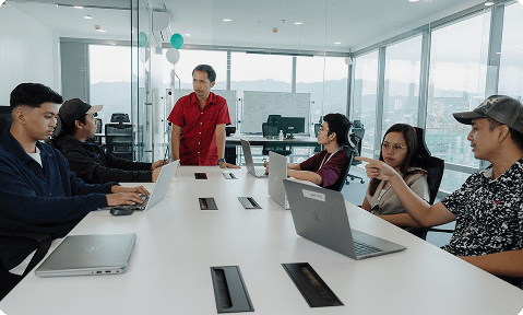 Six people are having a meeting in a glass-walled office; four are seated with laptops, one is standing and speaking, and another gestures while listening.