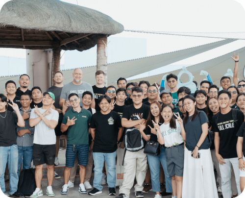 A diverse group of people posing together outdoors, some smiling and holding gestures, under a thatched roof with a clear sky in the background.