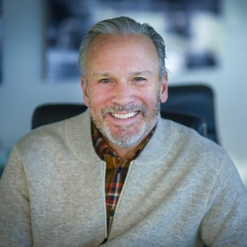 Portrait of a smiling elderly man with a beard, wearing a sweater and plaid shirt, seated in an office chair.