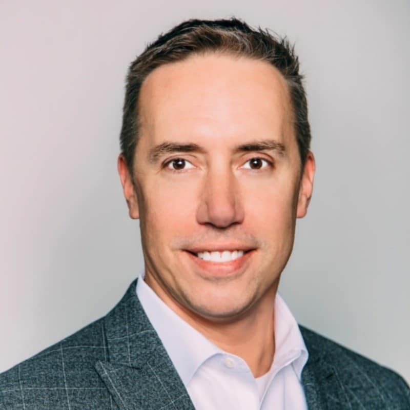 A professional headshot of a smiling man with short brown hair, wearing a gray blazer and a light blue shirt.