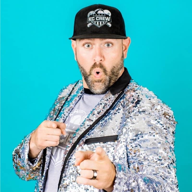 A man with a beard, wearing a shiny silver jacket and a black cap, looks surprised while pointing directly at the camera against a turquoise background.