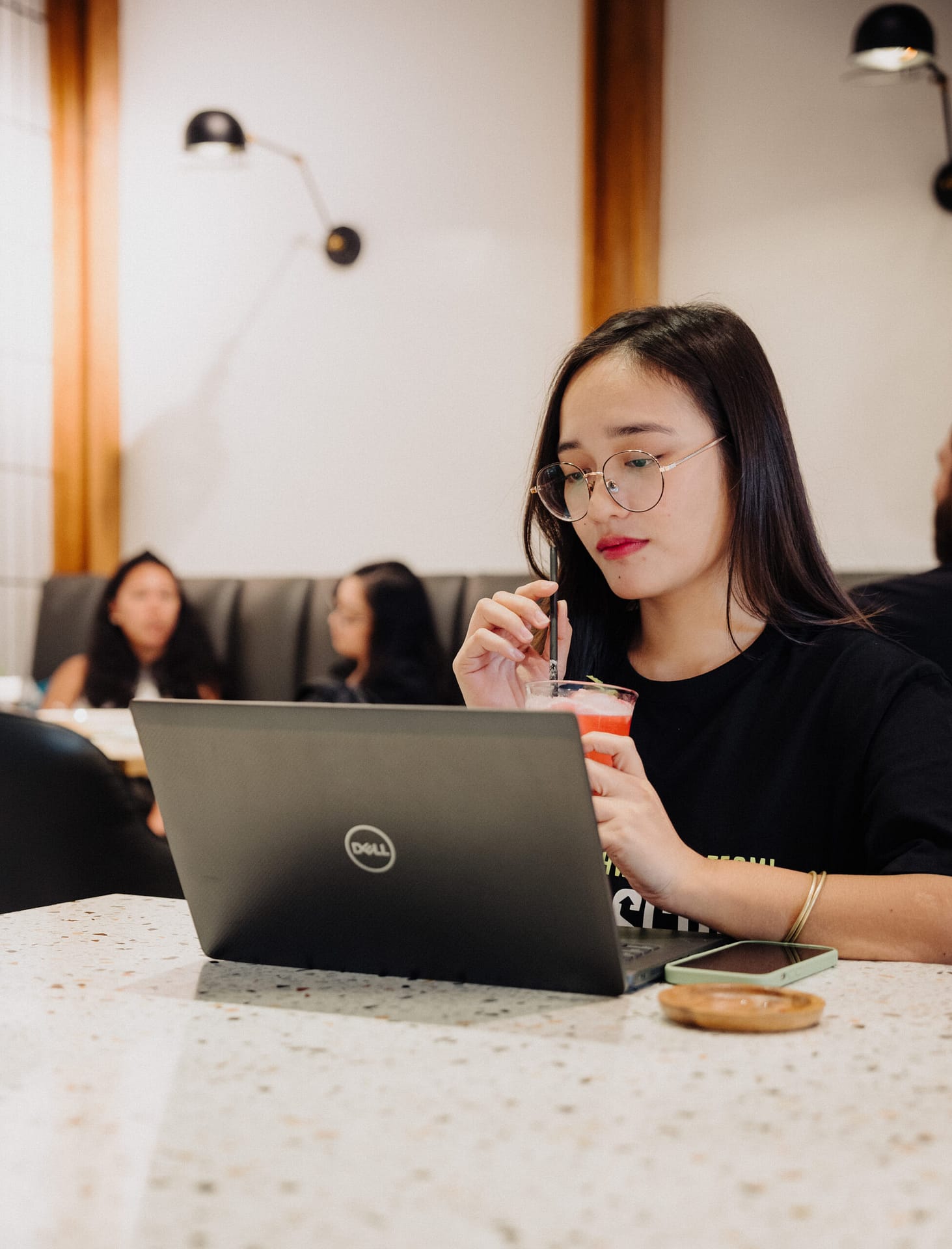 A woman with glasses works on a Dell laptop at a cafรฉ, sipping a drink through a straw. Other people are seated and talking in the background.