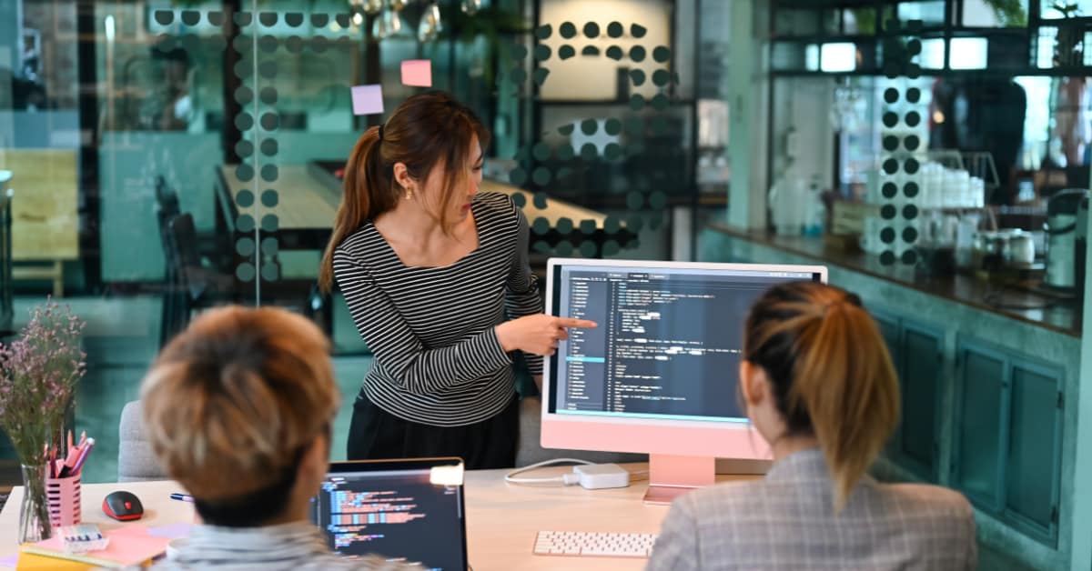 A woman in a striped top is presenting code on a computer screen to software testers in an office setting.