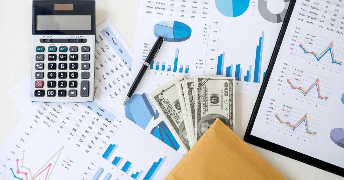 A financial analysis setup with a calculator, various financial graphs and charts printed on paper, a pile of US dollar bills, and a brown envelope on a white surface.