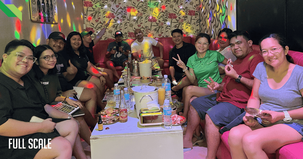A full scale team sits on red sofas around a table with food and drinks, basking in colorful lights, smiling for a photo.