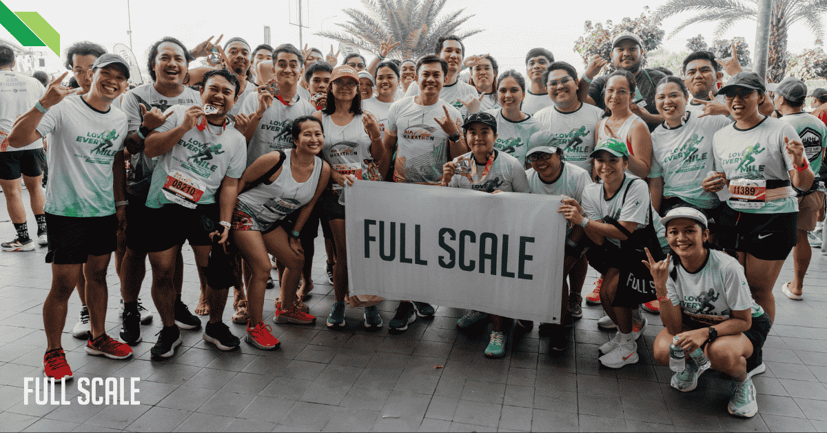 A group of people in running attire pose with a banner reading "FULL SCALE" at an event.