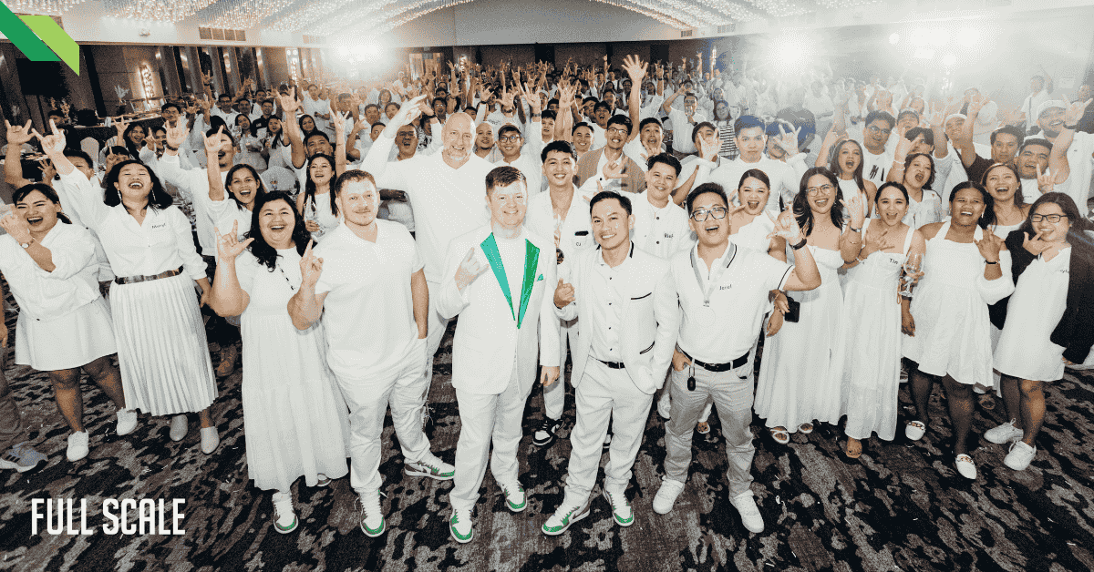 A large group of people dressed in white posing for a photo in a well-lit event space. Some are making peace signs. "Full Scale" is visible in the bottom left corner.