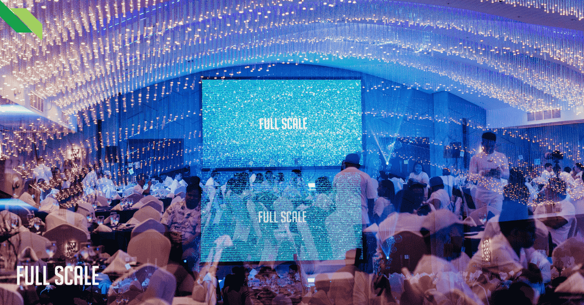 A crowded event hall with people in white attire, a large screen displaying "FULL SCALE," and ceiling lights creating a twinkling effect.