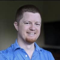 A man in a blue shirt, representing an offshore development company, smiles in front of a TV.