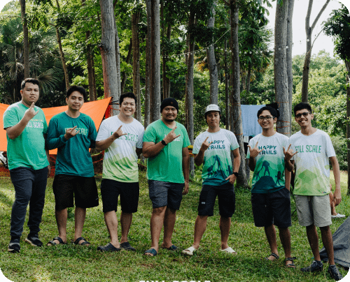 Seven people standing on grass in a forested area, wearing casual summer clothes, some with graphical t-shirts. Orange tents and trees are visible in the background.