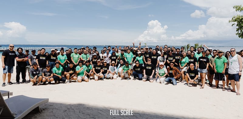 A large group of people posing for a photo on a beach, with many wearing green and black "Full Scale" T-shirts. The ocean and a cloudy sky are in the background.